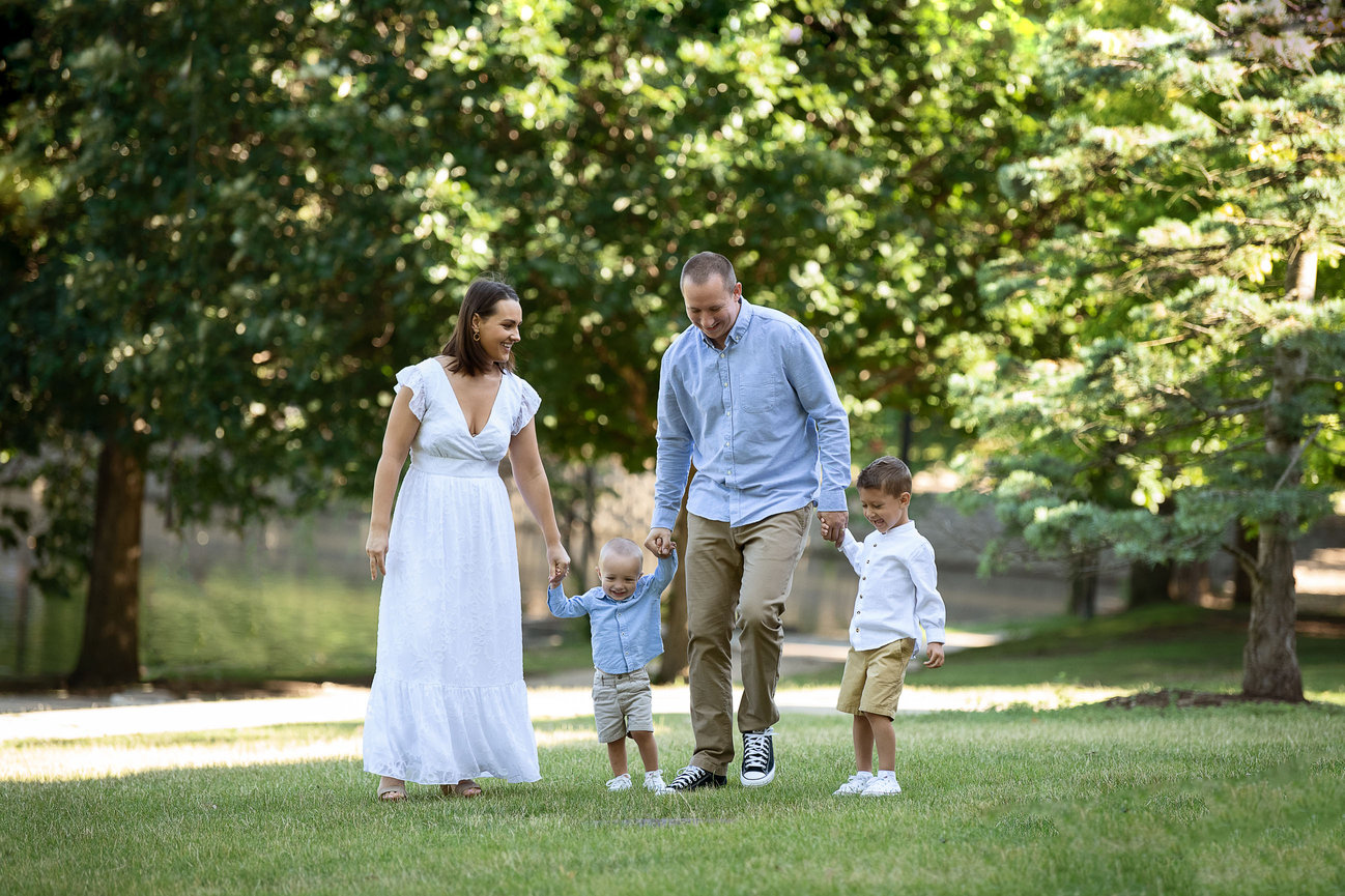 A family of four strolls hand in hand through Lords Park, Illinois. The woman wears a white dress, the man in a blue shirt and khaki pants, and their two young kids are dressed in blue and white shirts with shorts. Lush green trees form the backdrop.