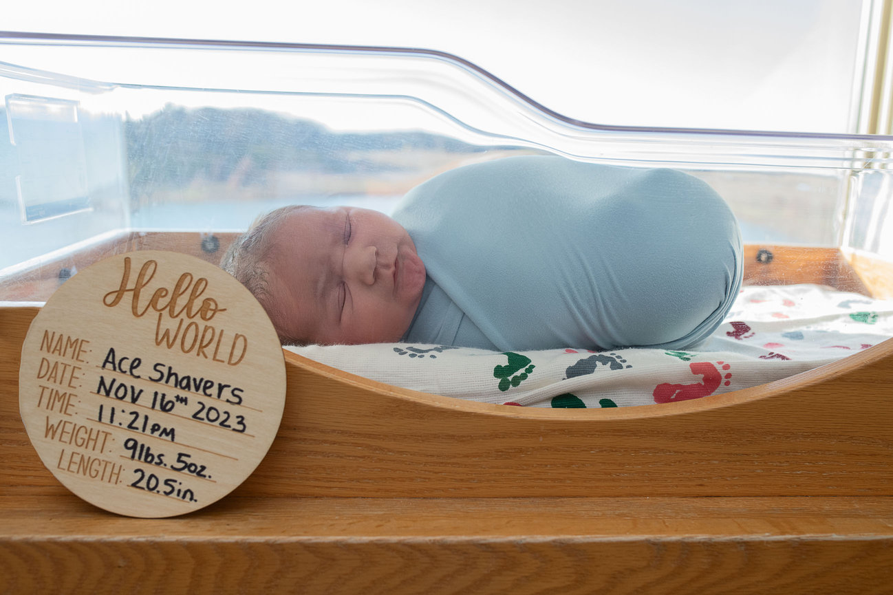 A newborn baby swaddled in a blue blanket sleeps peacefully in an Elgin hospital bassinet. A wooden sign beside the baby reads: “Hello World. Name: Ace Shavers. Date: Nov 16th, 2023. Time: 11:21 PM. Weight: 9 lbs. 5 oz. Length: 20.5 in.”.