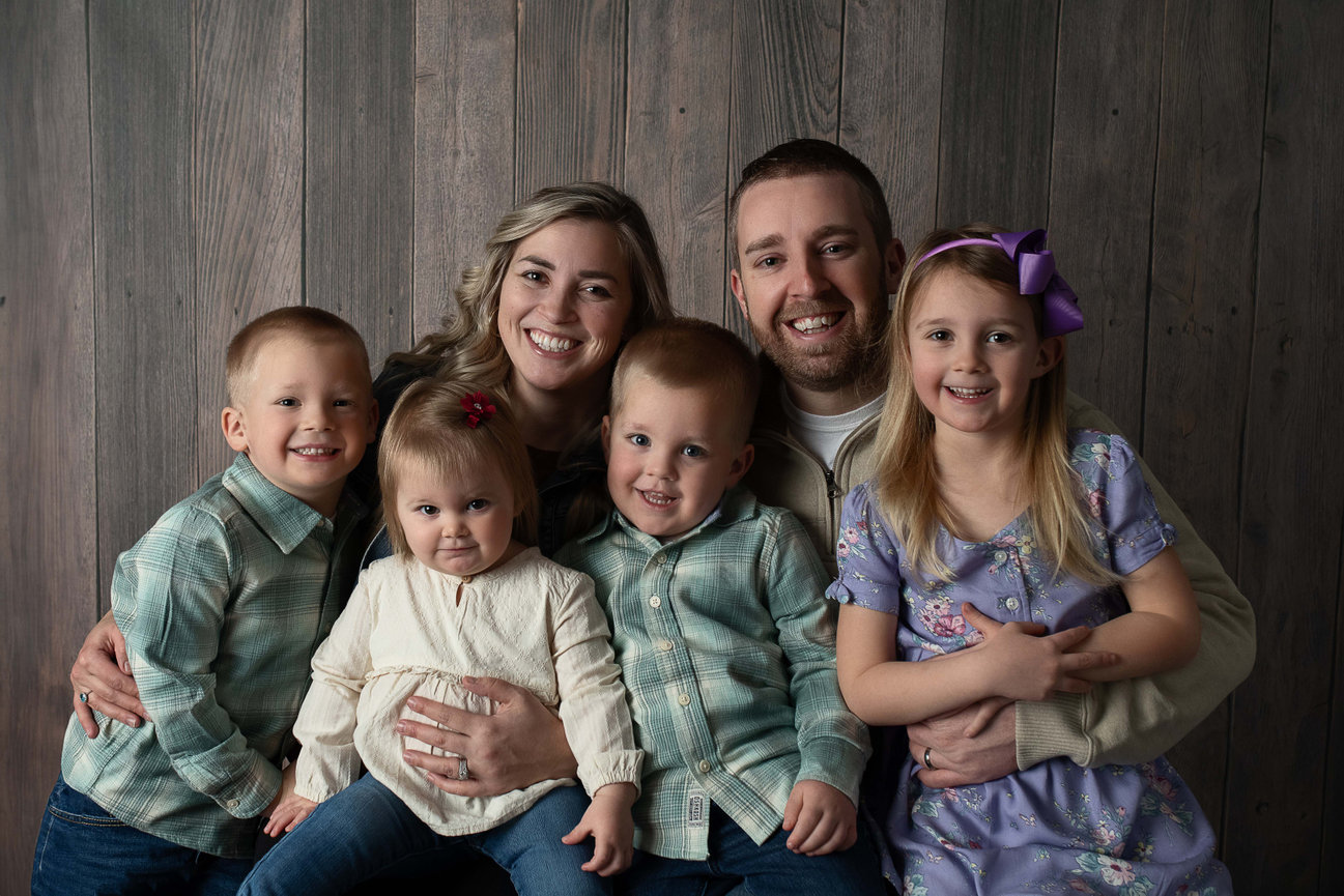 A smiling family poses together in front of a wooden wall, capturing a moment with the expertise of a studio family photographer. The group includes two adults and four young children, casually dressed, with one girl with a purple bow in her hair.