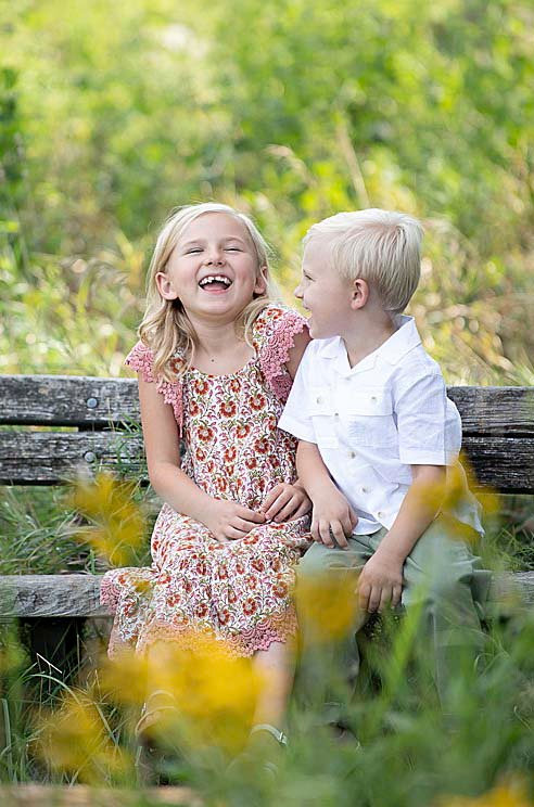 Two young children sitting on a wooden bench amidst lush greenery delight in each others company. The girl, in a floral dress, joyfully laughs while the boy, wearing a shirt, smiles at her. A perfect moment captured by a family photographer near me.