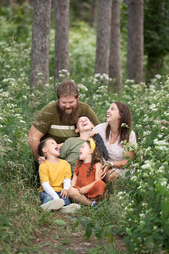 A family sits together in a lush, green forest, captured perfectly by a skilled family photographer. A bearded man and a woman are seated behind three children, all laughing joyfully. The trees and wildflowers around them in Lake in the Hills.