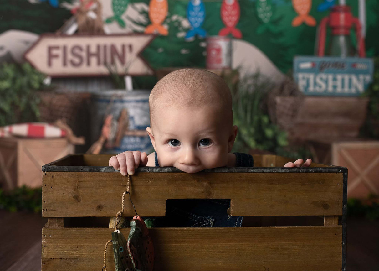 A baby peeks over the edge of a wooden crate, set against a backdrop featuring fishing-themed decor, including signs and colorful fish illustrations. The baby is holding a toy fish.