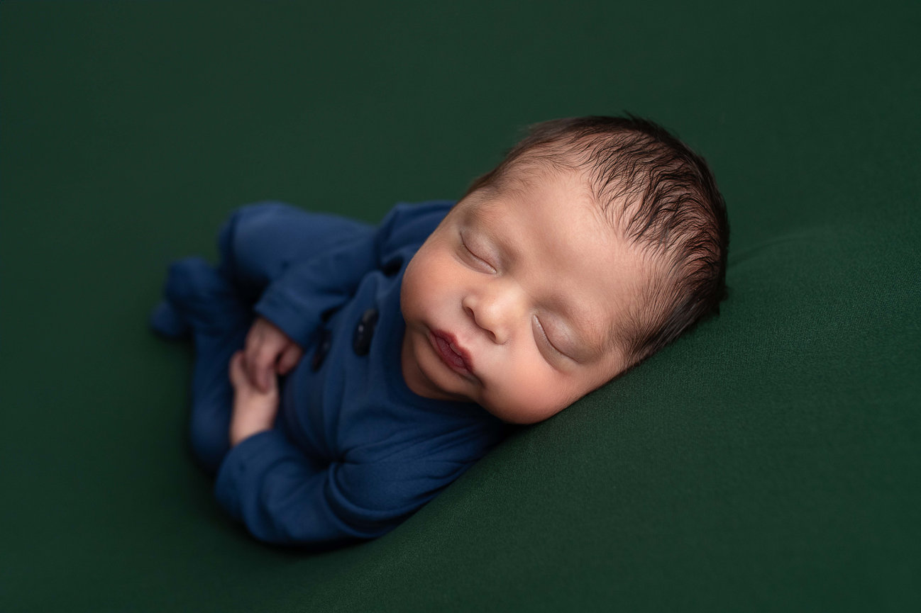 A newborn baby peacefully sleeping on a dark green surface, wearing a blue outfit. The babys hands are gently clasped in front, and they have dark hair. The serene scene evokes the tranquility of an Illinois wildrose bloom.