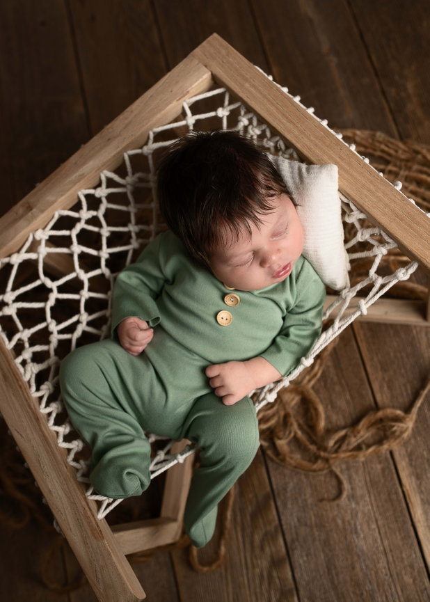 A sleeping baby in a green onesie rests in a wooden-framed net hammock, with a matching green pillow under their head. The serene scene unfolds on a wooden floor in St. Charles, Illinois, complemented by a soft, brown textured blanket underneath.