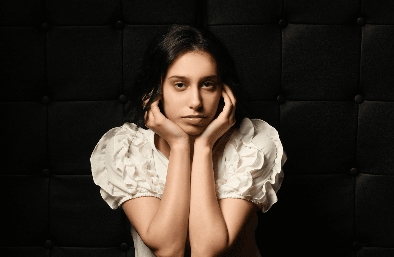 A high school senior gal wearing a white frilly sleeved blouse resting her chin in her hands.