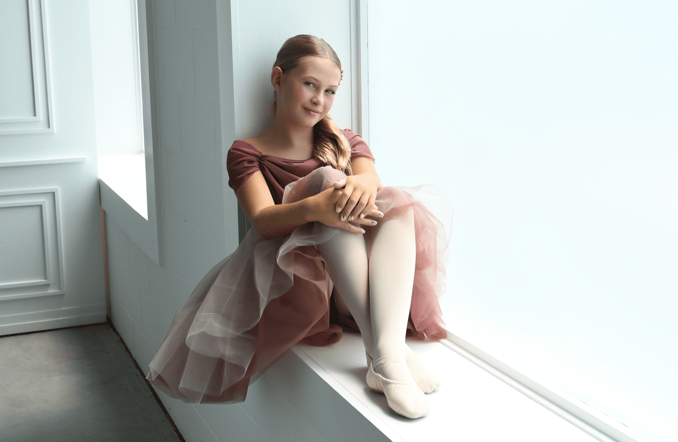 A pre-teen girl sits in the window sill wearing her pink ballet dress an slippers for a dance session at Kliks Photography.