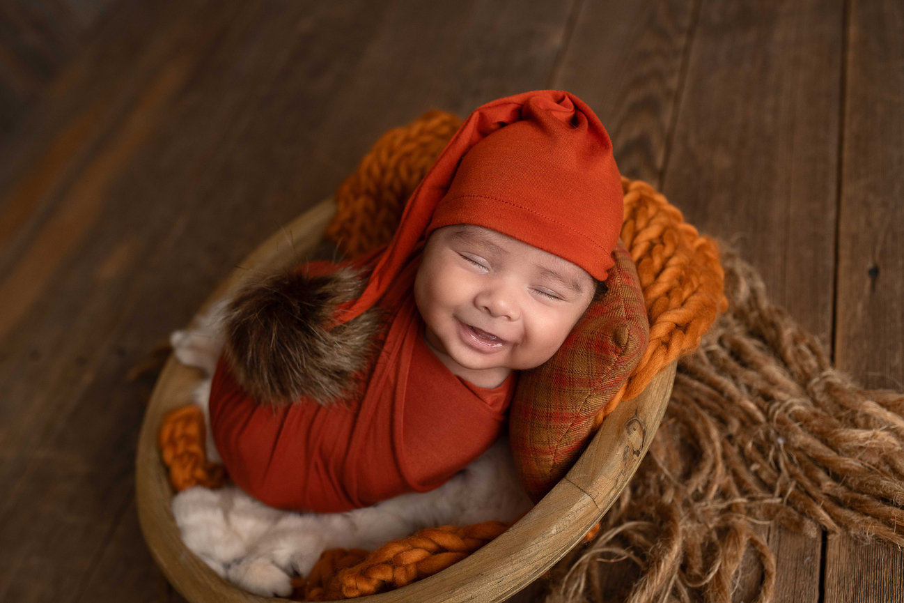 Smiling newborn baby dressed in a cozy orange swaddle and matching hat, lying in a rustic wooden bowl with soft textures – professional newborn photography in Barrington, Illinois