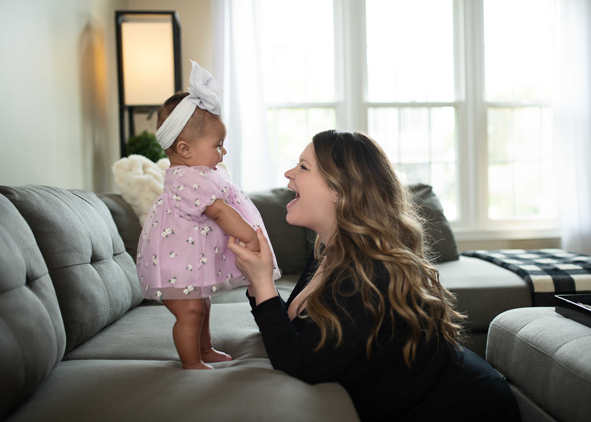 Mother smiling at baby girl standing on couch during in-home lifestyle photography session in natural light