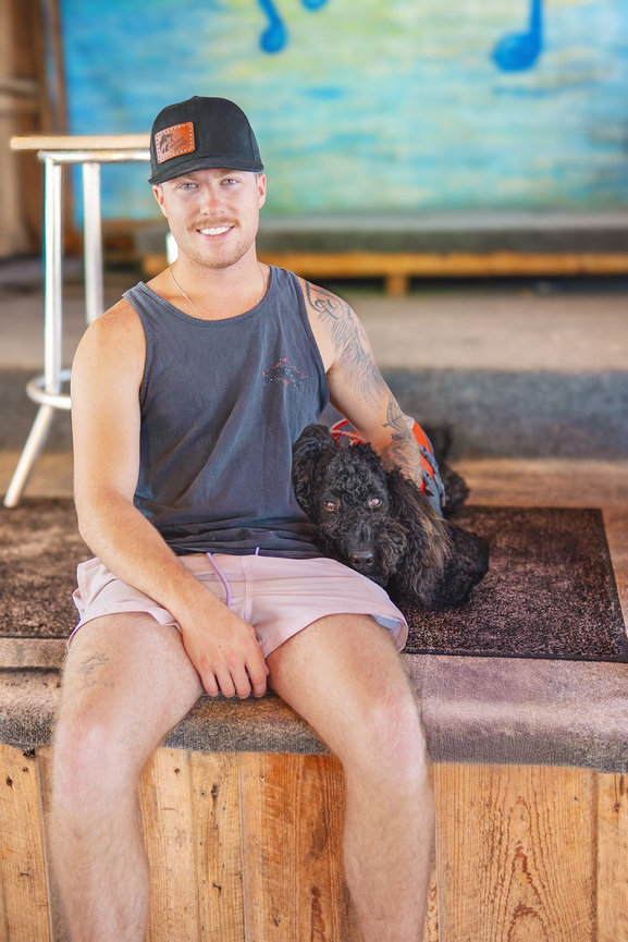Man in a black cap and tank top sitting on a wooden stage with a black dog resting on his lap.