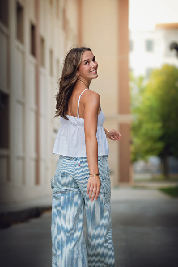 High School senior girl in white top and striped pants walking outdoors, smiling, with blurred buildings and greenery in the background in downtown kenosha.