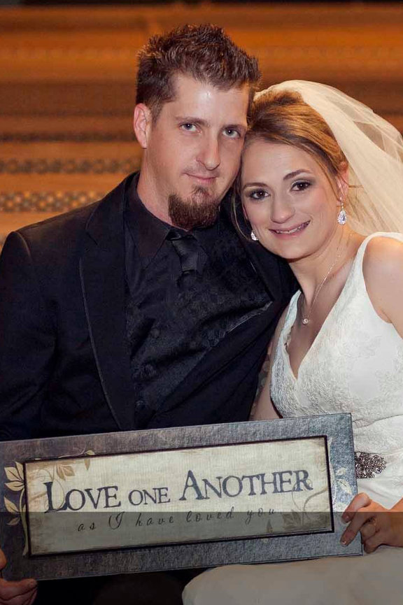 Anna & Blaine sit closely holding a sign that says Love one Another as I have loved you right after their wedding, with PBM Photography.