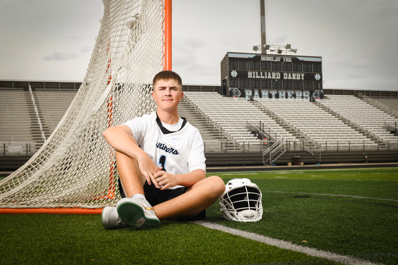 High school lacrosse player sitting by the goal, wearing a jersey and helmet on the field.