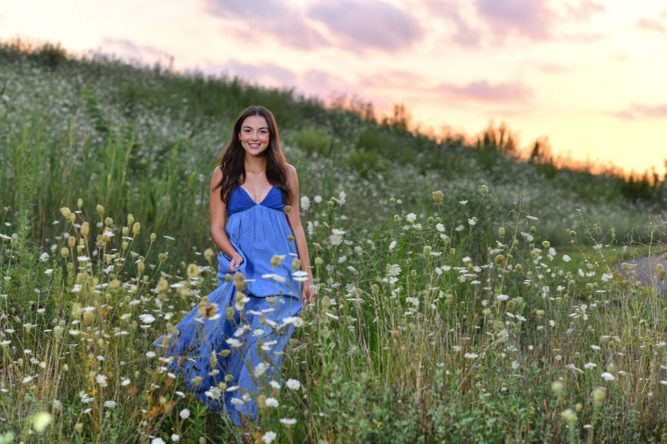 Teen girl in a flowing blue dress stands in a field of wildflowers at sunset for senior pictures in Columbus Ohio.
