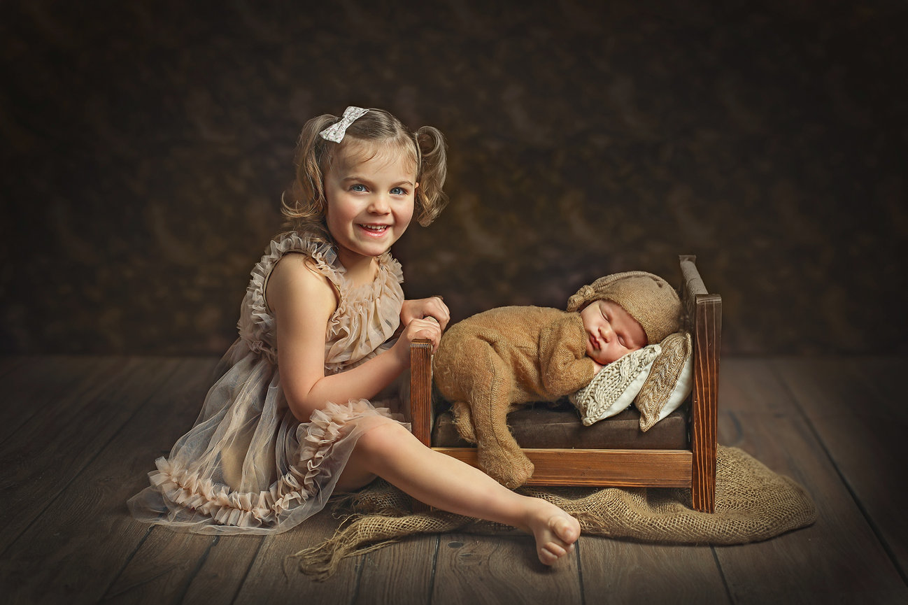 Young girl smiling beside a newborn baby sleeping in a small wooden bed, with both wearing brown outfits on a wooden floor.