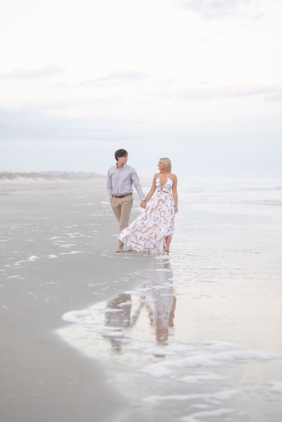 Couple holding hands and walking barefoot on a beach, with their reflection in the wet sand.
