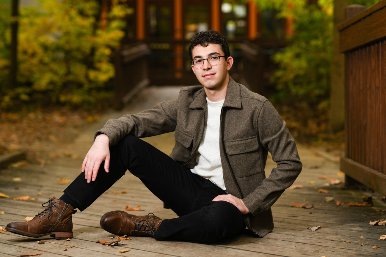 Dark-haired teenage boy in black jeans and a brown jacket sitting on a rustic bridge with yellow and orange fall leaves around him.