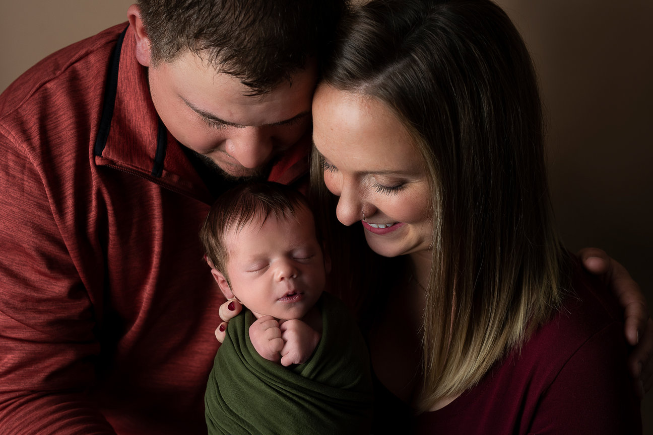 In Lake Barrington, Illinois, a couple lovingly embraces their sleeping baby, wrapped in a green cloth. The parents, wearing maroon and brown tops, smile gently as they gaze down at the infant against a warm, neutral background.